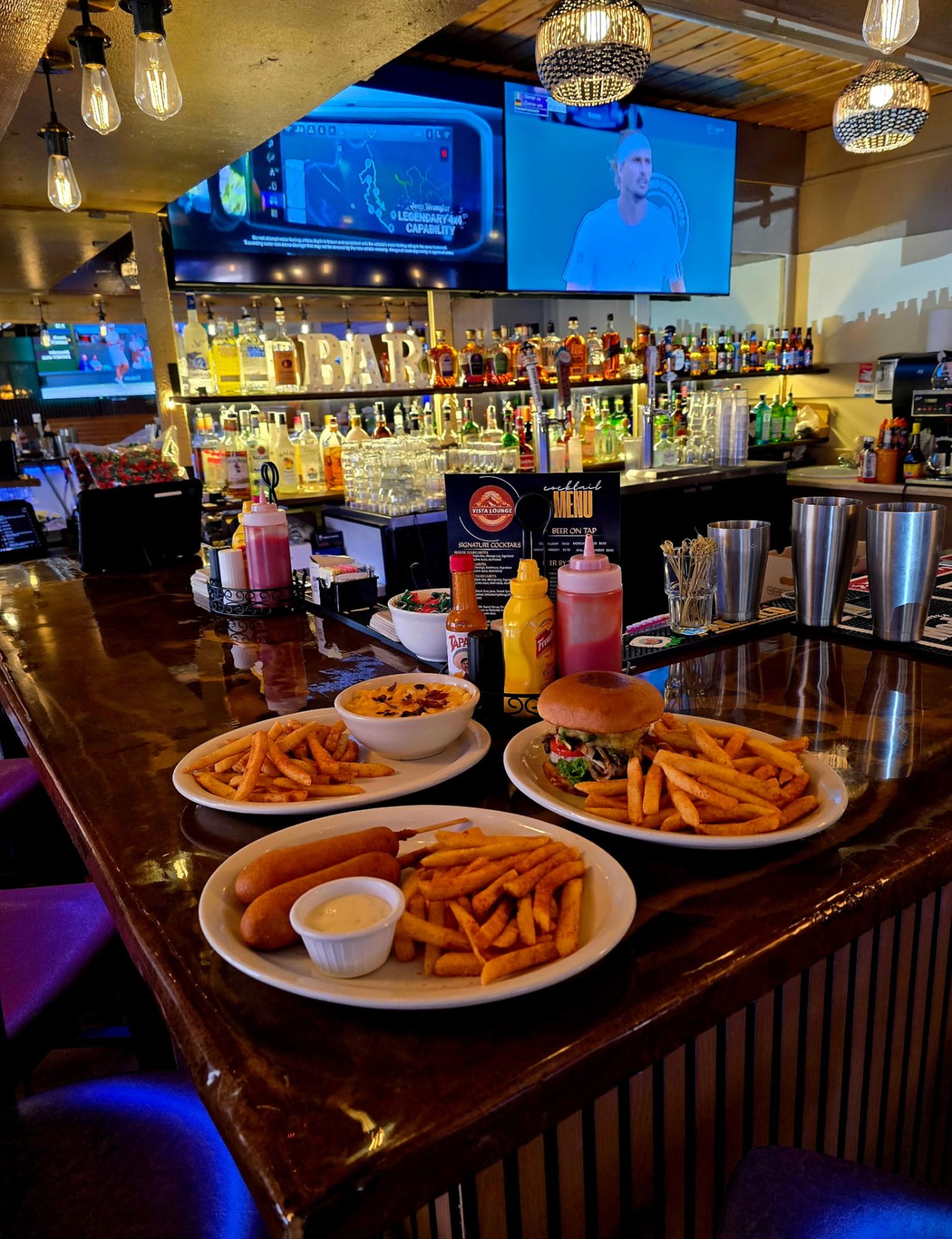A mushroom Swiss burger with fries, a bowl of loaded mac and cheese and a corn dog basket lined up on the Vista Lounge bar