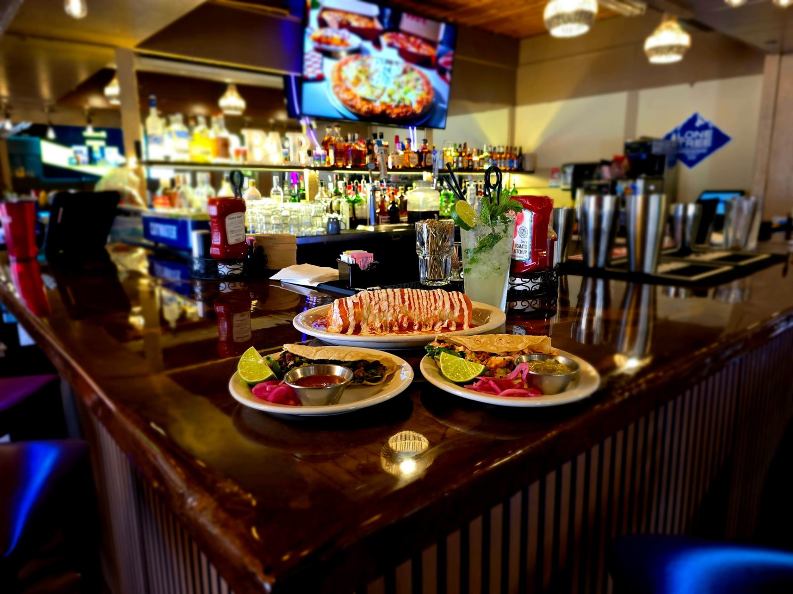 A smothered burrito and two plates of tacos on the polished wood bar under Edison bulb lights